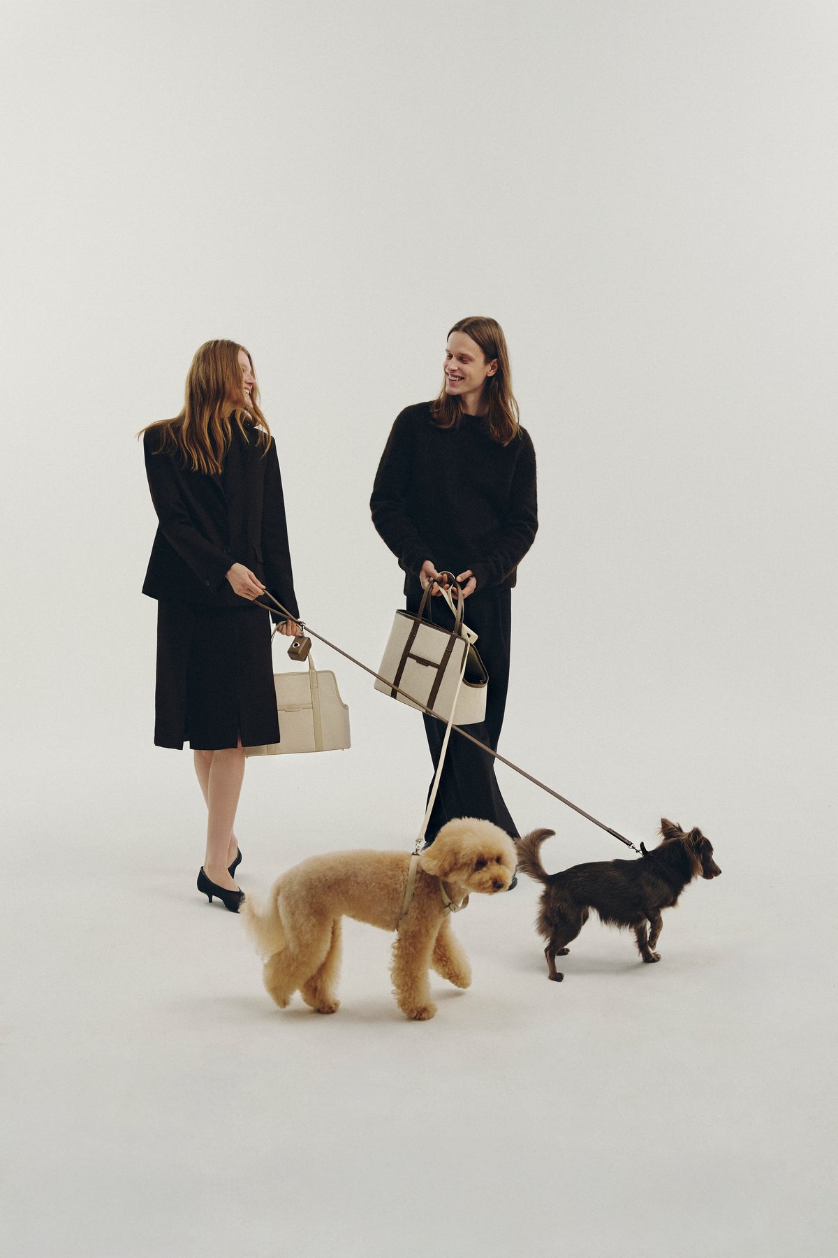 Two women in black outfits pose with two small dogs and carry cream-colored structured handbags in a minimalist studio setting.