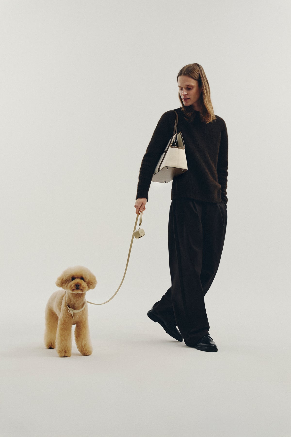 A woman in a black long-sleeve top and wide-leg trousers walks with a small beige poodle on a cream leash, carrying a white shoulder bag against a plain light gray studio background.